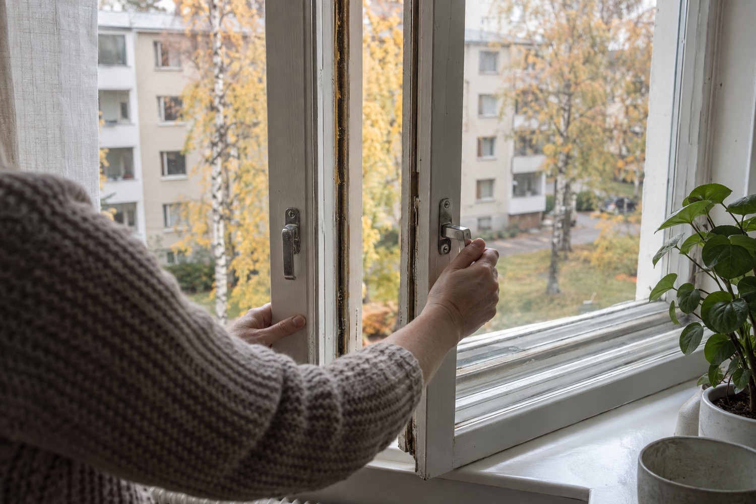 Henkilö avaa kerrostaloasunnon kaksipuitteisen ikkunan sisäpuitetta syksyn päivänvalossa